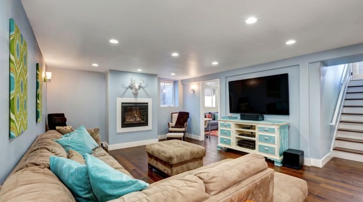 Inviting basement living area with soft blue walls, dark hardwood floors, a gas fireplace, large sectional sofa with turquoise pillows, wall-mounted TV on distressed console, and decorative art—showcasing full finishing and ambiance.