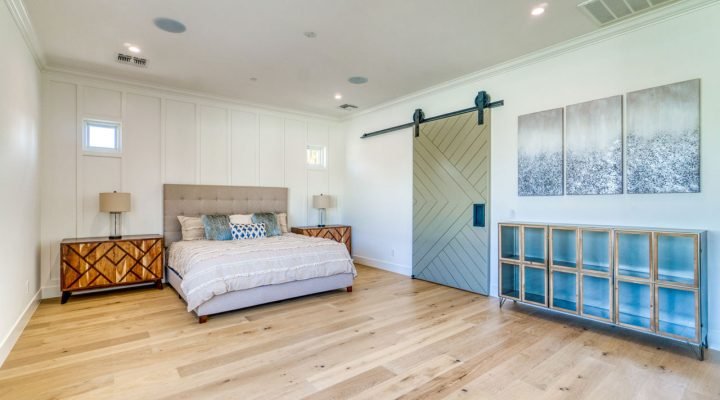 Bright, modern bedroom featuring light oak hardwood flooring, white shiplap walls, a tufted bed with patterned bedding, geometric wood nightstands, a gray barn door on black track, and a blue-glass cabinet—showcasing Teton Repair & Remodel’s interior finishing expertise.